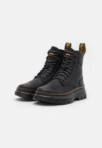 Bottes de combat en cuir noir à lacets avec semelles épaisses à rainures et coutures jaunes, avec des languettes de tirage marquées à l’arrière.