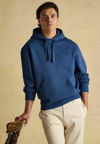 Tume sinine kapuutsiga särk, puuvilla segu, esipocketiga, soonikuga varrukatega ja logoga kapuutsil. Modell poseerib tooliga.