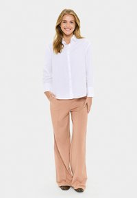 Smiling woman with long blonde hair wearing a white button-up shirt and beige wide-leg pants, standing with hands in pockets.