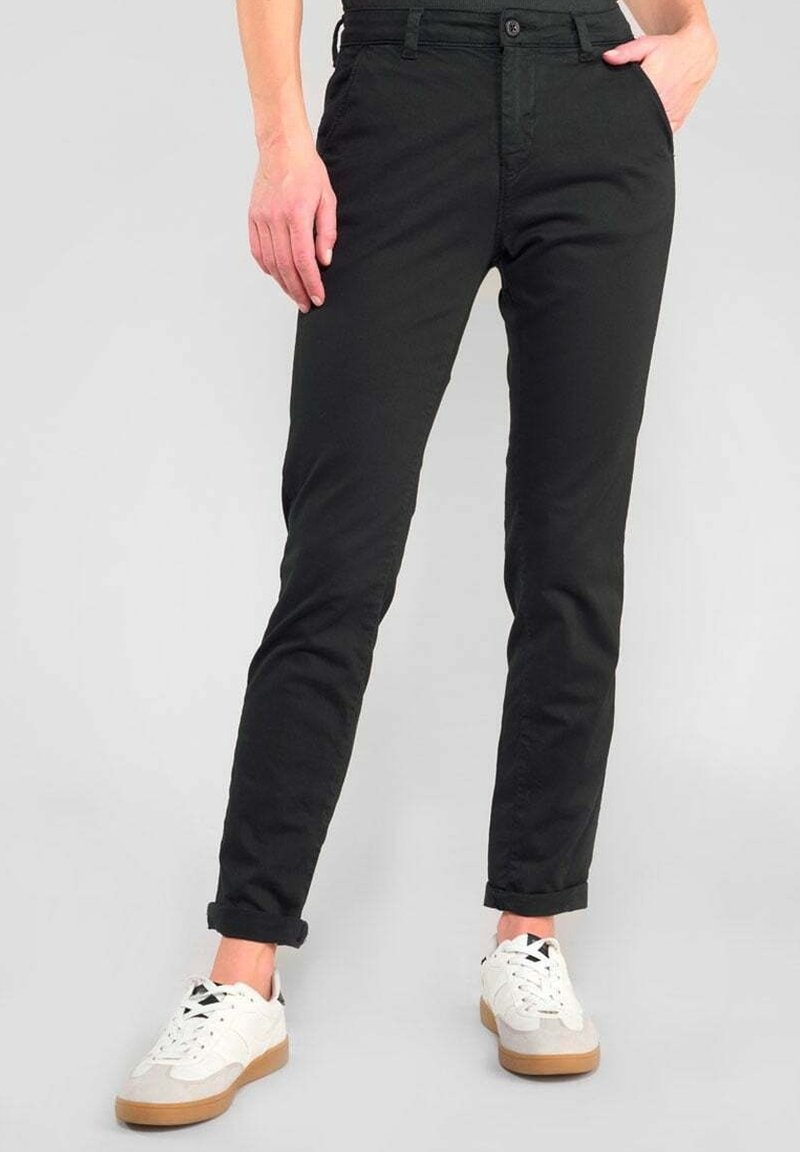 Personne portant un pantalon noir ajusté et des baskets blanches, debout avec une main dans la poche sur un fond gris.