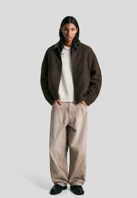 Rjav zip-up jopič z zelenim ovratnikom, bel pleten pulover, bež kordne hlače in črne čevlje. Model stoji z rokami v žepih.
