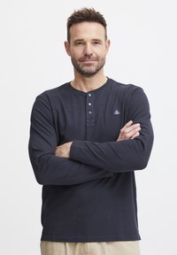 Marineblå langermet henley skjorte med tre knapper ved kragen, strukturert stoff og en liten logo-patch på venstre bryst.