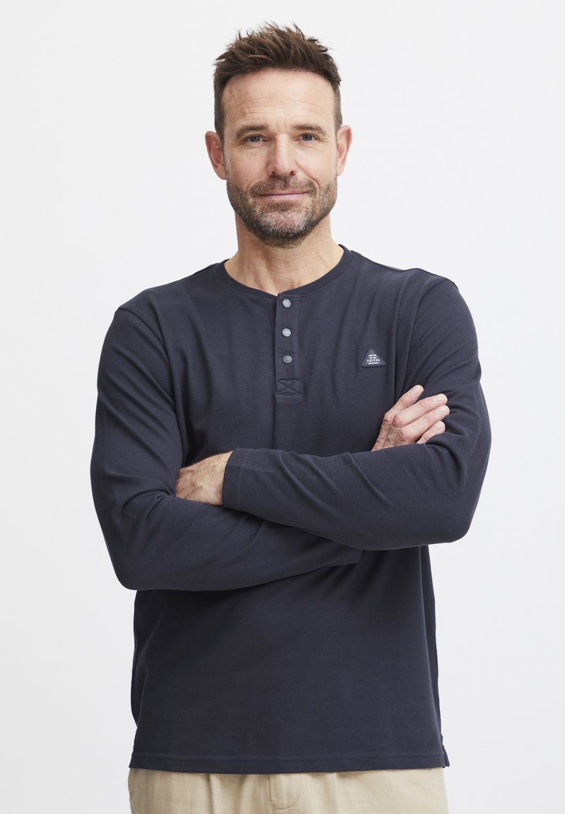 Marineblå langermet henley skjorte med tre knapper ved kragen, strukturert stoff og en liten logo-patch på venstre bryst.