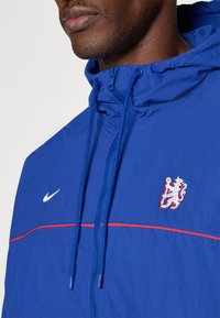 Blå nylonjakke med hette som kan lukkes med glidelås, røde detaljer, hvit Nike-logo og et hvitt brodert løveemblem på brystet.