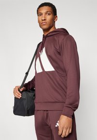 Sweatshirt marrom com um logótipo branco, apresentando um capuz com cordão, mangas longas e tecido texturizado. Acompanhado de uma bolsa preta.