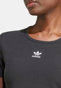 Juodas raukšlėtas marškinėlis su apvaliu kaklu ir trumpomis rankovėmis; krūtinėje yra baltas Adidas logotipas. Minkšta audinio tekstūra.