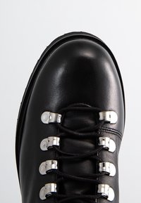 Botte en cuir noir à bout arrondi avec des lacets noirs maintenus par six œillets en métal argenté sur fond blanc.