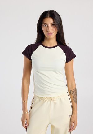 Jonge vrouw met lang donker haar, gekleed in een crème-zwart shirt met korte mouwen en crèmekleurige broek met donkere zijstrepen, staand tegen een effen achtergrond.