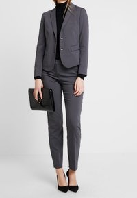 Veste grise ajustée avec pantalon slim assorti, un col roulé noir en dessous, et une pochette noire texturée. Des escarpins noirs complètent la tenue.