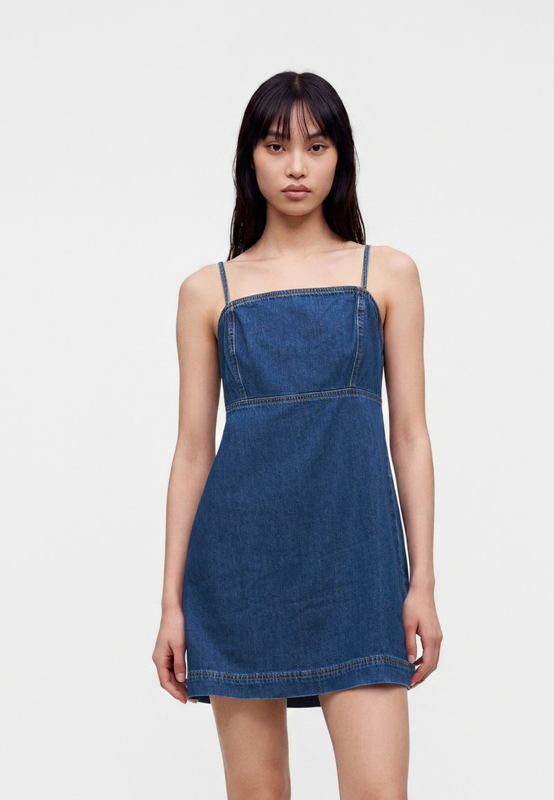 Jeune femme aux cheveux noirs et raides portant une robe en jean bleue sans manches avec des fines bretelles, sur un fond uni.