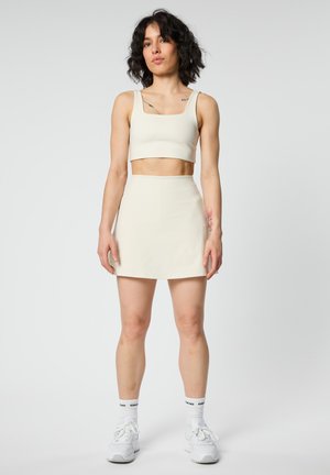 Rövid, göndör hajú nő, aki törtfehér crop topot, hozzáillő magasderekú szoknyát, fehér zoknit és fehér tornacipőt visel, egyszerű háttér előtt áll.