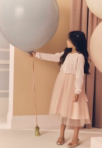 Abito rosa chiaro con sovrapposizione di tulle, cardigan bianco lavorato a maglia, mentre tiene un grande pallone grigio. Scarpe dorate e una base decorativa in oro per il pallone.
