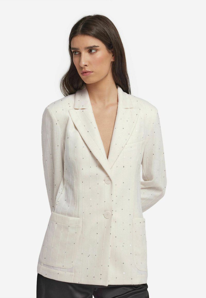 Biały żakiet z teksturowanej tkaniny, z pionowymi prążkami i srebrnymi cekinami. Wyróżniające się elementy to dwie kieszenie z przodu oraz kołnierz z wycięciem.
