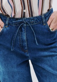 Pantalones de denim en azul oscuro con un corte holgado, que presentan una cintura con cordón, bolsillos frontales y detalles de costura visibles.