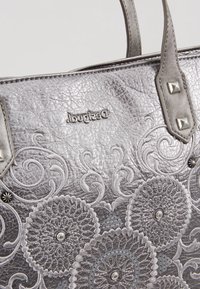 Srebrna skórzana torebka z wytłaczanymi kwiatowymi wzorami, ćwiekowanymi uchwytami oraz metalowym logo marki "Desigual" na przodzie.