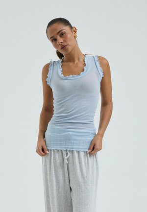 Žena s vlasmi stiahnutými dozadu, oblečená v svetlomodrom tielku bez rukávov s volánovými okrajmi a voľných svetlosivých vzorovaných nohaviciach, stojaca pred jednofarebným pozadím.