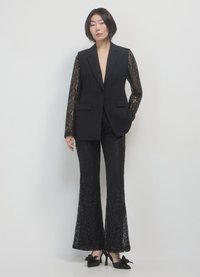 Giacca nera con maniche lunghe in pizzo, abbinata a pantaloni neri a palazzo in pizzo. Scarpe nere con tacco e dettagli a fiocco completano l'outfit.
