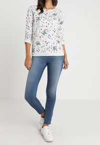 Sudadera floral gris claro con patrones en azul, rojo y verde, combinada con jeans ajustados azules y zapatillas blancas.