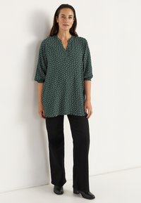 Groene tuniek met een patroon, een V-hals en 3/4 mouwen, gecombineerd met zwarte rechtlopende broeken en zwarte laarzen met hak.