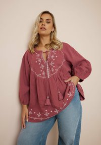 Dieprode gemarmerde blouse met geborduurde details, puffed sleeves en franjes, gecombineerd met losvallende blauwe jeans.