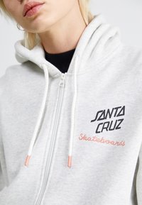 Haut de survêtement zippé gris clair avec capuche à cordon. Présente des logos brodés en noir et rose "SANTA CRUZ" et "Skateboards". Matériau en coton.
