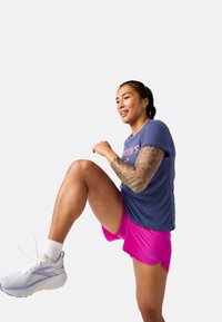 Atleta con una camiseta azul y pantalones cortos rosas, realizando un ejercicio de rodillas altas. Sus características incluyen un tatuaje en su brazo izquierdo y zapatillas deportivas blancas.