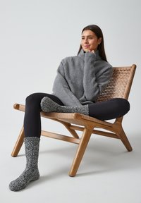 Suéter gris de canalé oversize, leggings negros y calcetines texturizados en blanco y negro, usados mientras está sentado en una silla de madera con asiento de mimbre.