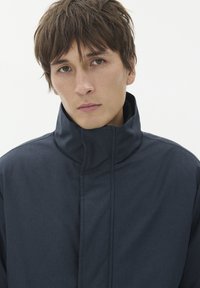 Jauns vīrietis ar īsiem brūniem matiem, ģērbies tumši zila augsta apkakles žaketē, skatās tieši uz priekšu ar neitrālu izteiksmi uz vienkārša balta fona.