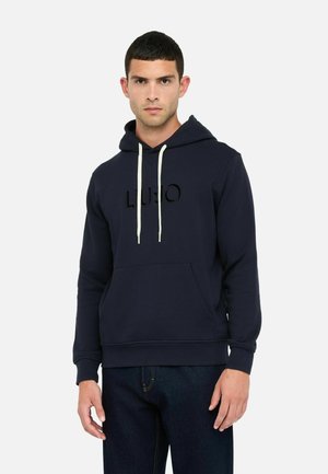 Mand iført en marineblå hættetrøje med hvide snore og frontlomme, parret med mørkeblå jeans, stående mod en hvid baggrund.