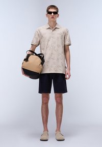 Beige pikétröja med löv mönster, marinblå shorts och beige slip-in skor. Bärande en rund tan och svart väska, kompletterad med mörka solglasögon.