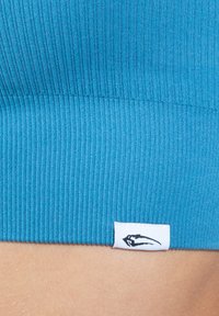 Tela de ribete en color azul verdoso, vista en primer plano, con una etiqueta blanca que contiene un logo negro, mostrando textura y elasticidad, con un acabado sin costuras.