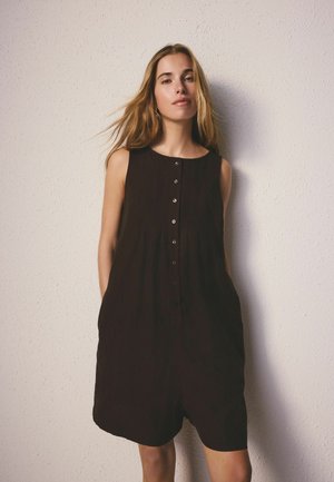 Młoda kobieta o długich blond włosach nosi czarną sukienkę bez rękawów na guziki, stoi oparta o jasną teksturowaną ścianę z rękami w kieszeniach.