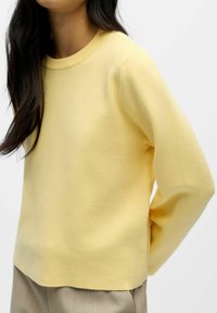 Kollane pika varrukaga sviiter, millel on soonikuga ümar kaelus, pehme puuvilla tekstuur, mugav lõige ja sirge äär, kantud beežide pükste peal.