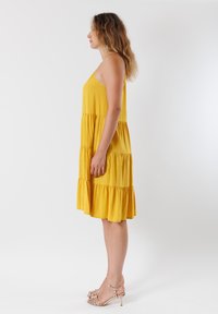 Abito giallo a strati con spalline sottili, realizzato in tessuto leggero. Fino sopra il ginocchio, con orlo arricciato. Indossato con tacchi a strappo.