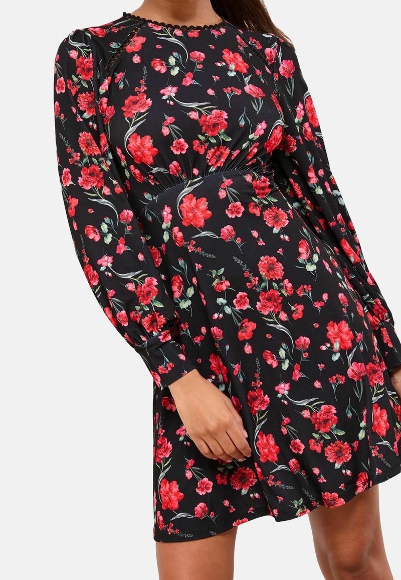 Robe noire avec imprimé floral rouge et rose, manches longues bouffantes, taille élastique et bordure en dentelle autour de l'encolure portée par une femme.
