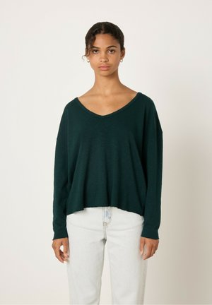 Top verde oscuro, holgado, de manga larga y con un amplio escote en V. La tela ligera y el dobladillo relajado complementan unos jeans de color claro.