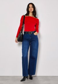 Rotes Off-Shoulder-Oberteil mit drapiertem Design, kombiniert mit hochtaillierten blauen weit geschnittenen Jeans und schwarzen Absatzstiefeletten. Schwarze Schultertasche.