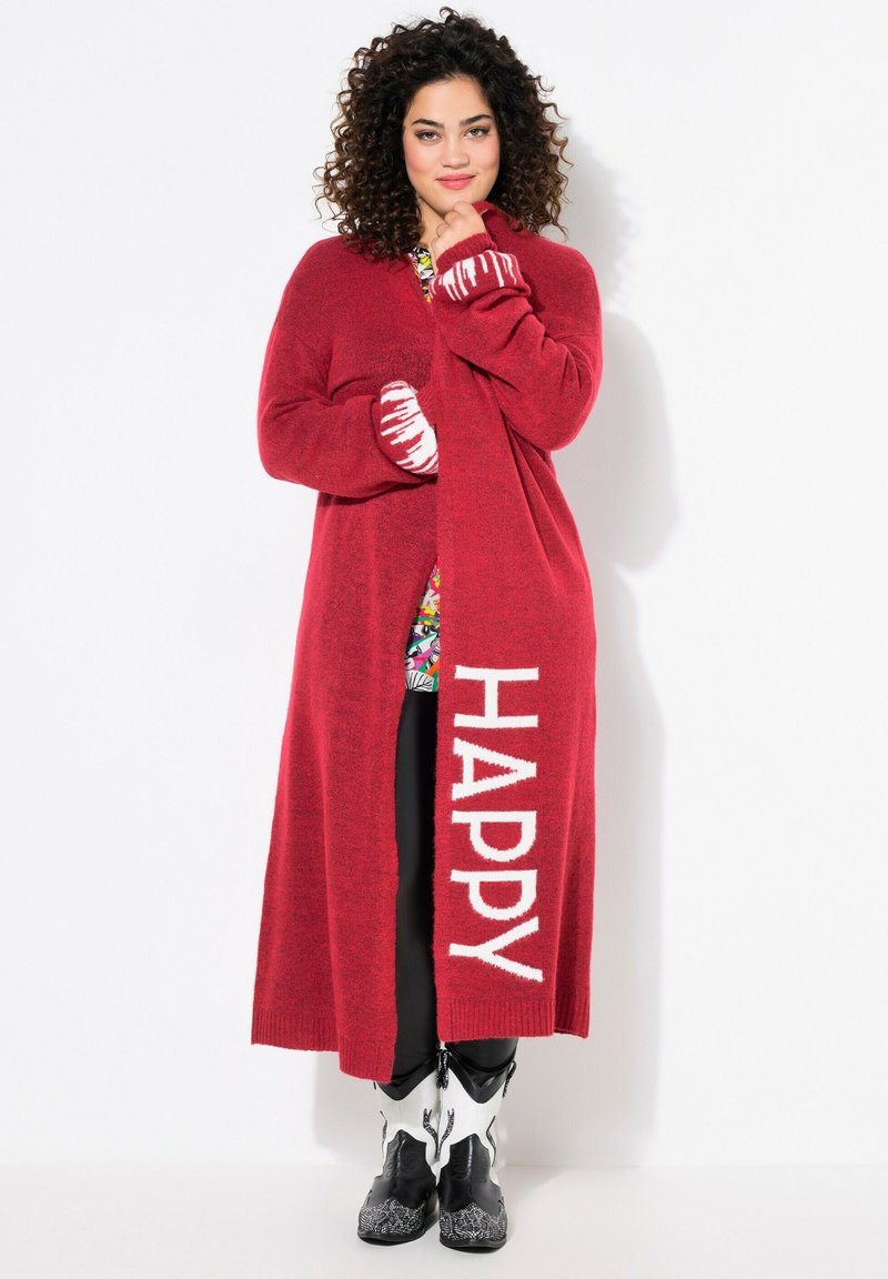 Cardigan long rouge avec le texte "HAPPY" en blanc. Matériau texturé et poignets rayés. Associé à un pantalon noir et un haut à motifs en dessous.