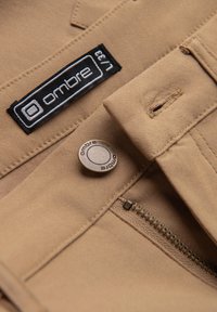 Gros plan d’un pantalon beige avec braguette zippée, bouton en métal portant la mention « ombre » et étiquette de taille en tissu indiquant « L/33 ».