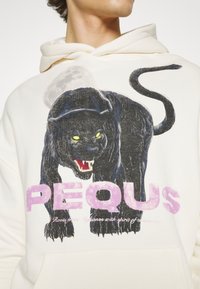 Cremefarbener Hoodie mit einem großen, schwarzen Panther-Grafik mit gelben Augen und geöffnetem Mund. Text "PEQUS" in Pink unter dem Design.