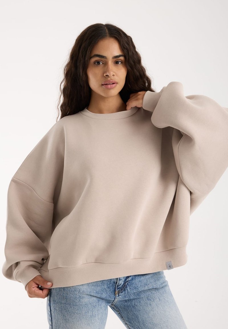 Przycinany beżowy sweter z ribbowanymi mankietami i dolnym wykończeniem, z okrągłym dekoltem i miękką teksturą, w połączeniu z jasnoniebieskimi dżinsami.