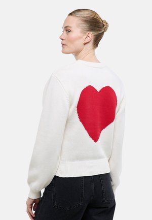 Femme aux cheveux blonds coiffés en chignon, portant un pull blanc avec un grand cœur rouge dans le dos et un jean noir, vue de profil.