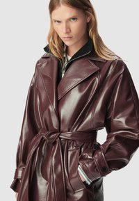 Person with long blonde hair wearing a shiny dark brown belted leather coat and a black zip-up layer underneath, hand in pocket.