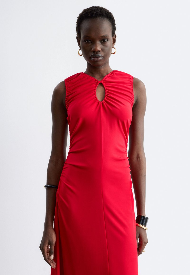 Femme portant une robe rouge sans manches avec un décolleté en trou de serrure et des détails froncés, debout devant un fond clair uni.