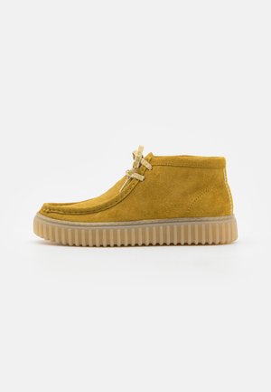 Botas de tobillo de ante amarillo con punta redonda, suela de goma beige, cordones en la parte delantera y parte superior texturizada.