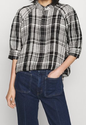 Femme portant un chemisier ample à carreaux noir et blanc avec des manches trois-quarts, rentré dans un jean taille haute bleu foncé avec des poches plaquées devant.