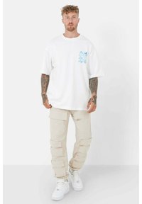 Vit bomullst-shirt med blå grafisk design på vänster bröst, tillsammans med beiga cargobyxor med sidofickor och utsvängda benslut, bärs med vita sneakers.