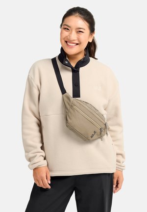 Lächelnde Frau, die einen beigen Fleecepullover und schwarze Hosen trägt, mit einer beigen Umhängetasche mit zwei Reißverschlusstaschen quer über der Brust.