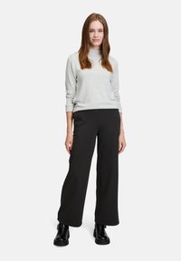 Grauer Pullover mit umgeschlagenem Kragen, kombiniert mit weit geschnittenen, karierter schwarzer Hose. Das Model trägt schwarze Stiefeletten und steht vor einem schlichten Hintergrund.
