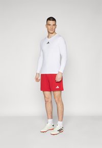 Chemise de sport à manches longues blanche avec un logo noir, associée à un short rouge arborant un logo, et des chaussures de sport blanches avec des accents noirs.